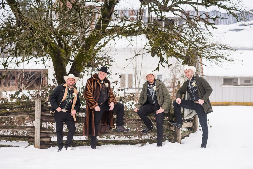 A groom and his three groomsmen, dressed in cowboy hats and boots, pose on a fence in the winter.