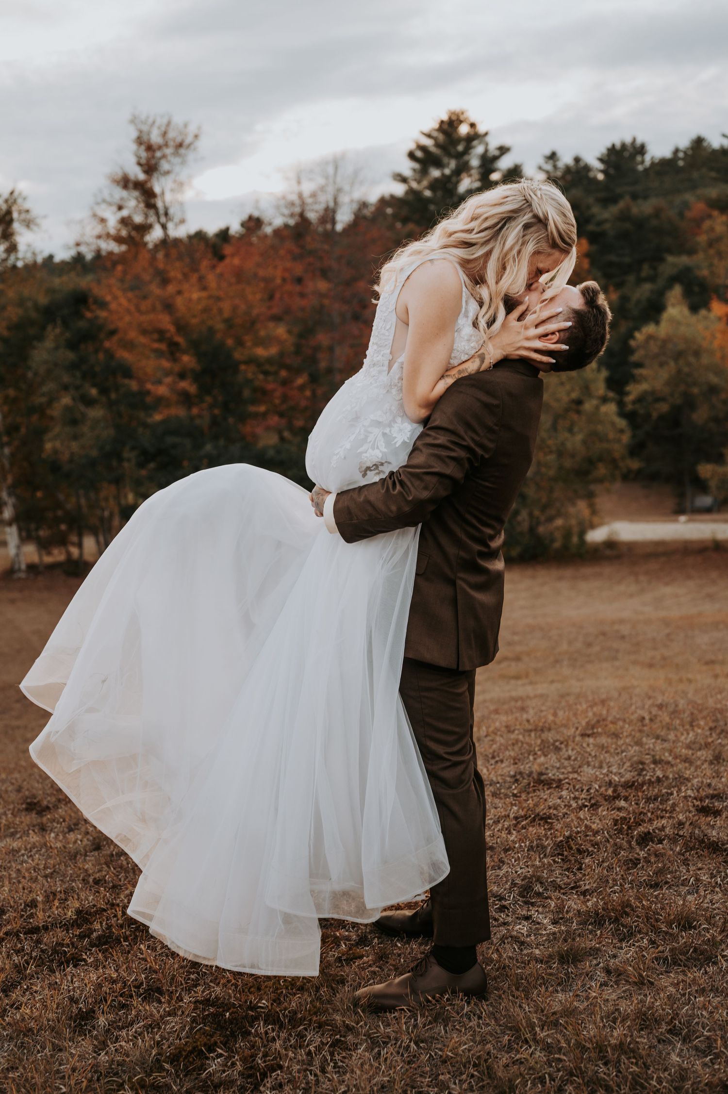 Groom lifting bride in white gown, sharing a romantic kiss outdoors with fall foliage in the background.