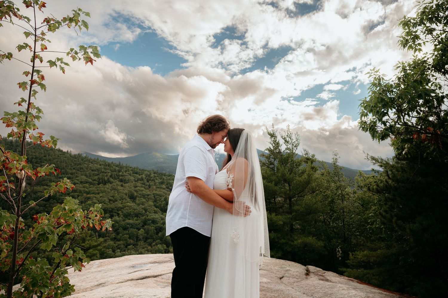 Groom lifting bride in white gown, sharing a romantic kiss outdoors with fall foliage in the background.