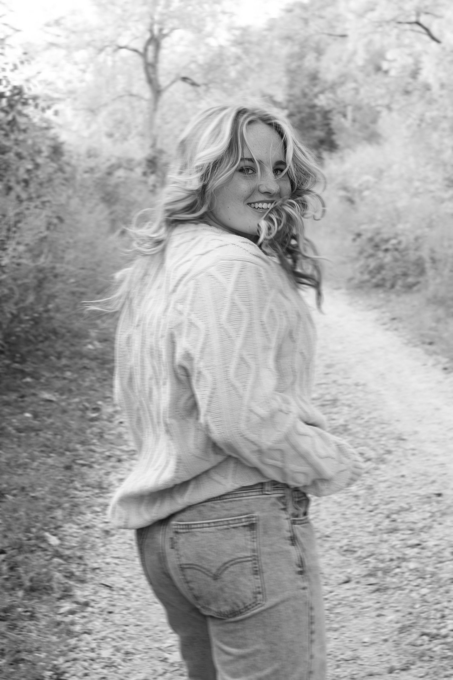 A black and white portrait of someone wearing a cable knit sweater and jeans on a forest path.