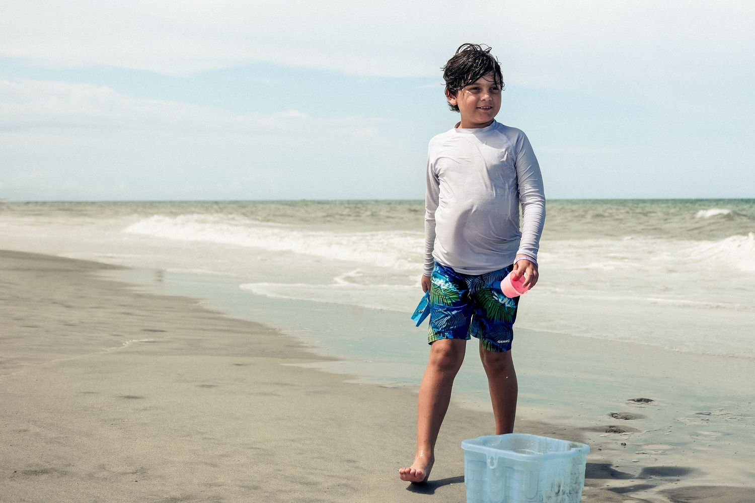 A child in beachwear stands on a sandy shore holding a blue bucket near the ocean waves.