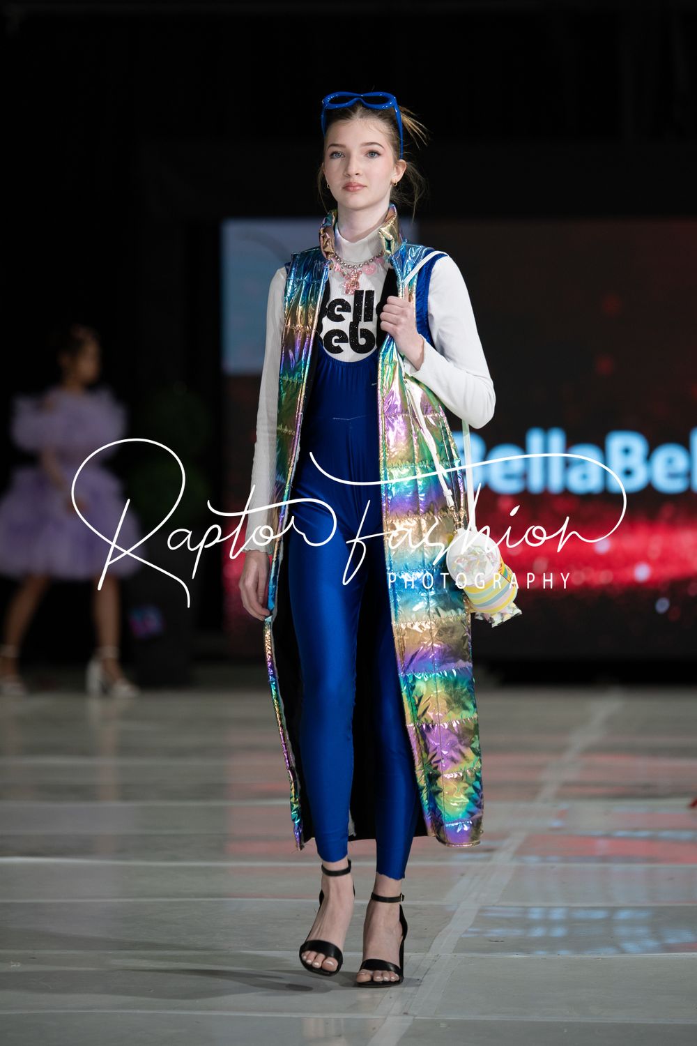 Model walks runway in navy blue jumpsuit with colorful floral kimono and blue headband at fashion show.