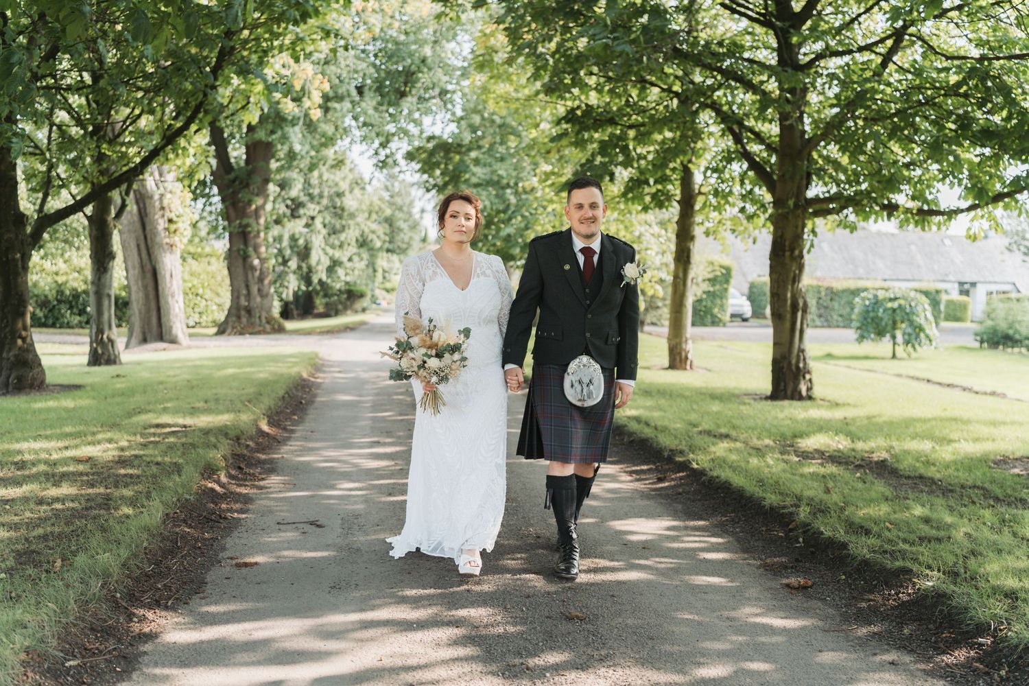 A couple walks arm in arm along a tree-lined path, with the groom wearing a traditional Scottish kilt and bride in white.