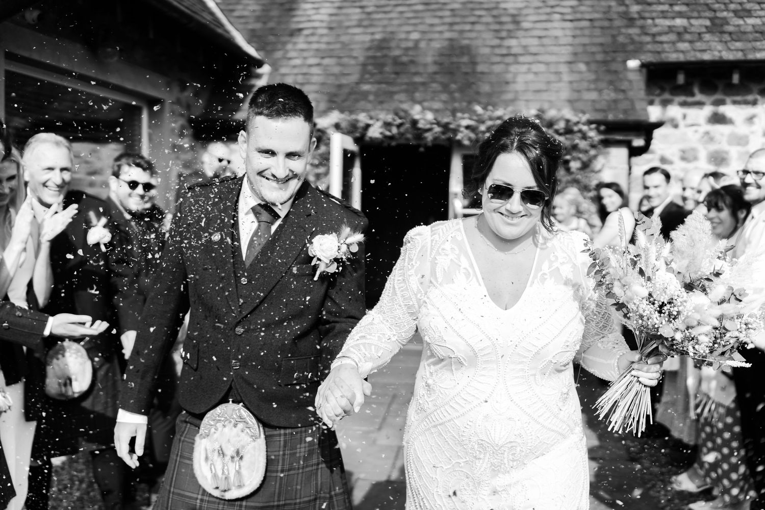 A joyful wedding couple walks together while guests throw confetti at their outdoor Scottish ceremony.
