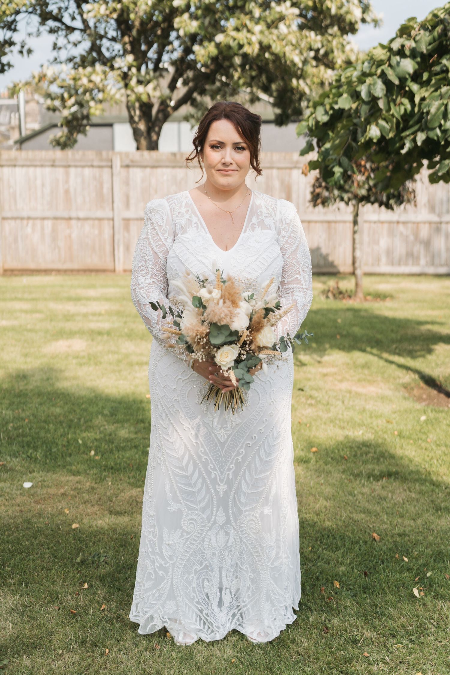A wedding image showing a white lace gown with long sleeves and a beautiful neutral-toned bouquet in an outdoor garden setting.
