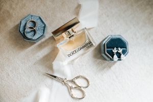 Dolce & Gabbana perfume, wedding rings, diamond earrings, and scissors arranged as bridal accessories.