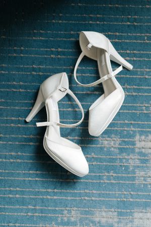 White satin bridal heels with ankle straps and rhinestone details on a teal textured carpet.