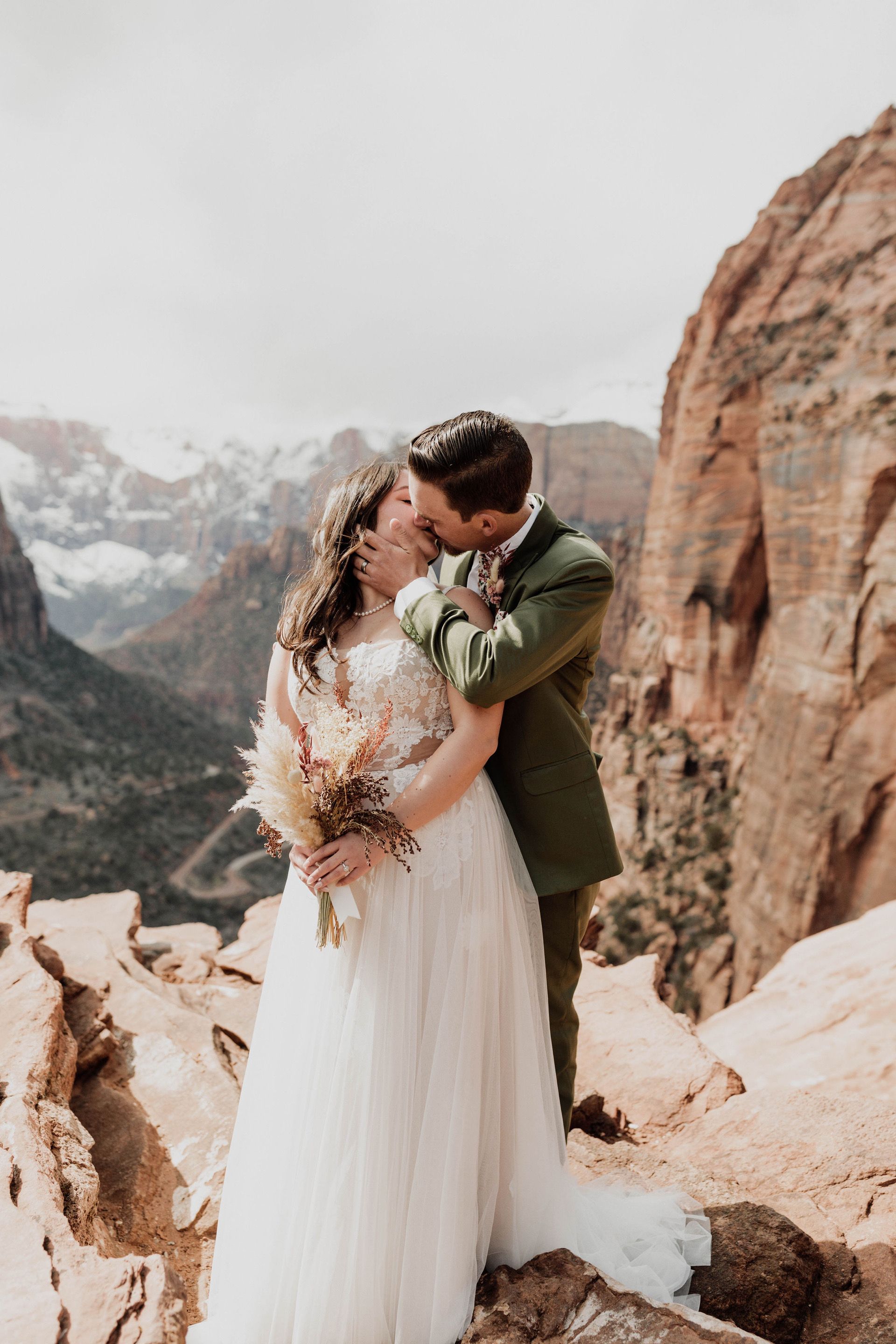 A couple shares an intimate moment against in Zion National Park's dramatic red rock cliffs and snow-capped mountains in the distance.