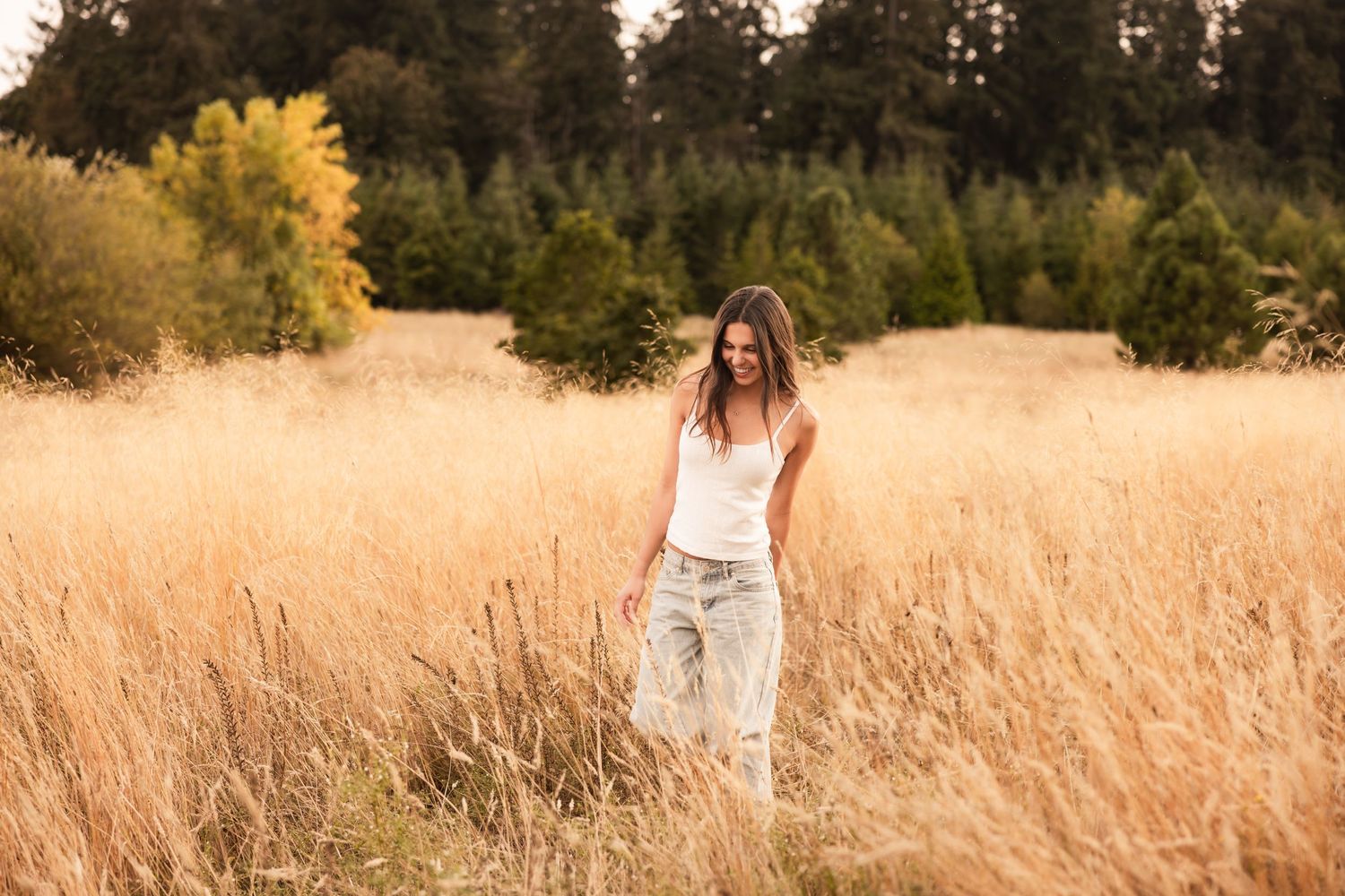 Person in white top and jeans standing in golden field of tall grass during sunset with trees in background.