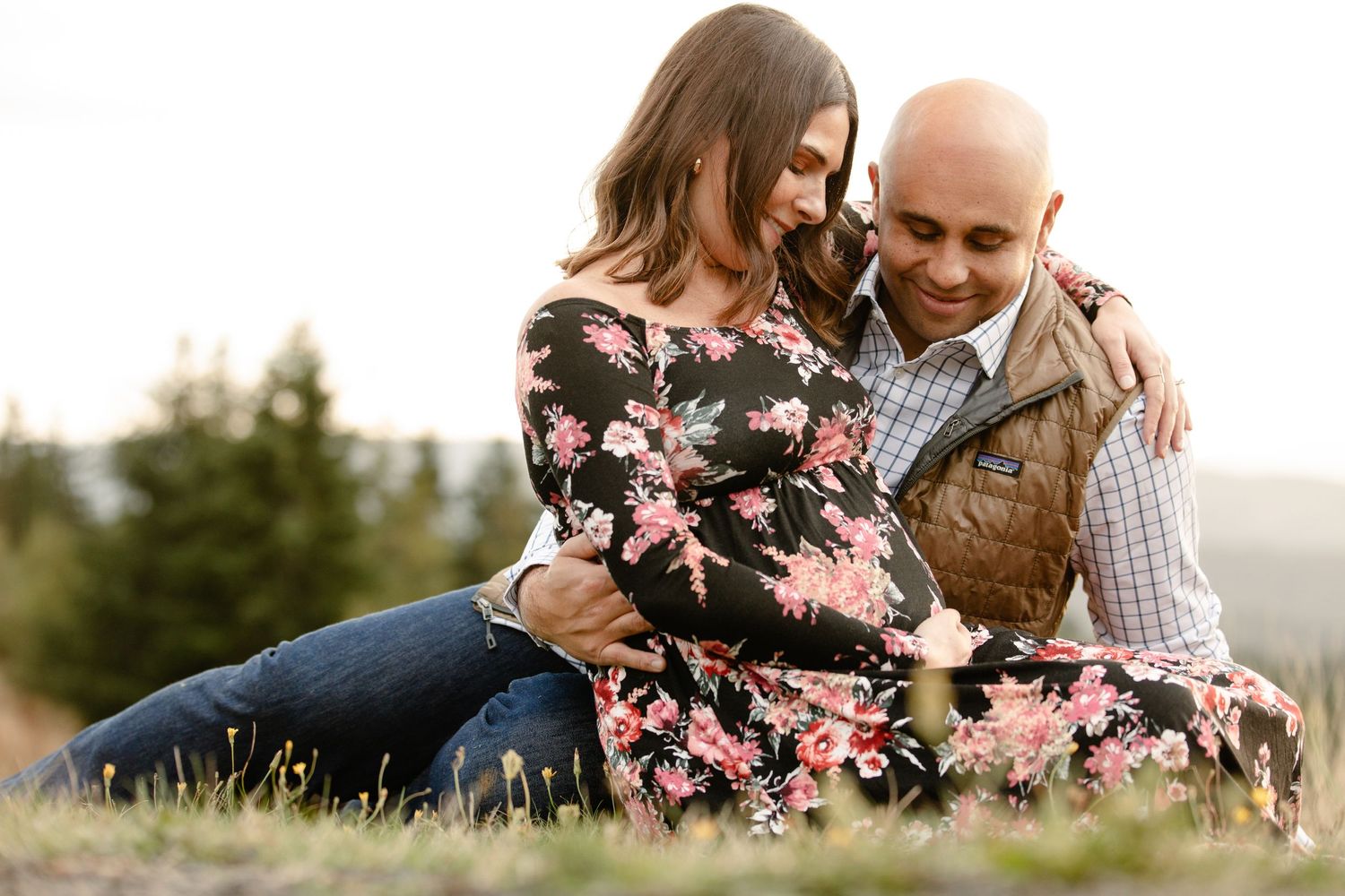 A couple sits together in a field at sunset, with her wearing a floral dress and him in a checkered shirt and vest.