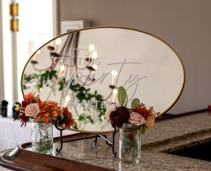 Elegant floral arrangements in mason jars next to an oval mirror on wooden table with golden trim.