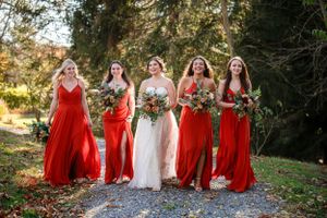 A bridal party in flowing red bridesmaid dresses and white wedding gown pose on a pathway surrounded by autumn foliage.