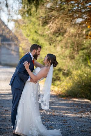 A romantic outdoor wedding photo of a couple embracing on a gravel path surrounded by autumn trees.