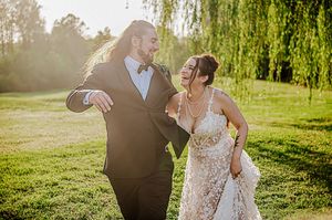 Joyful wedding couple dancing outdoors under golden sunlight and weeping willow trees.