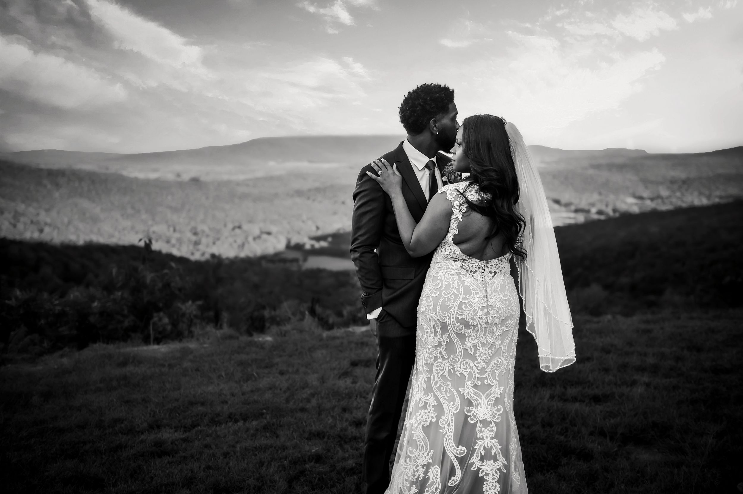 A black and white wedding photo of newlyweds embracing on a hilltop with mountain views.