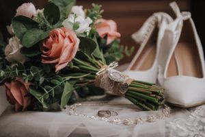 Wedding details including pink roses bouquet, white heels, and rings on delicate lace fabric.