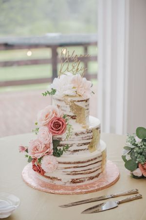 Semi-naked wedding cake decorated with pink roses and gold 'Better Together' topper on white table.