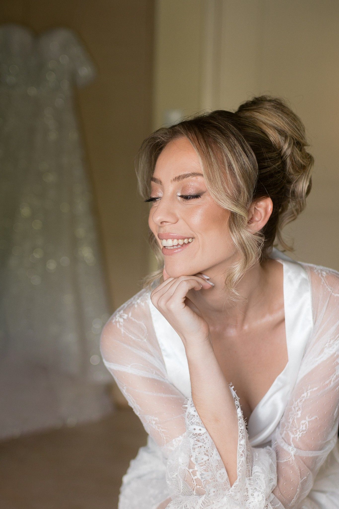 A bride in a white lace robe smiles joyfully while getting ready for her wedding day.