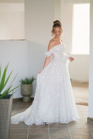 Bride in flowing white sparkly wedding dress twirls in bright sunlit room with potted plants.
