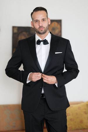 A person wearing a classic black tuxedo with bow tie buttons their jacket while standing in an elegant indoor setting.