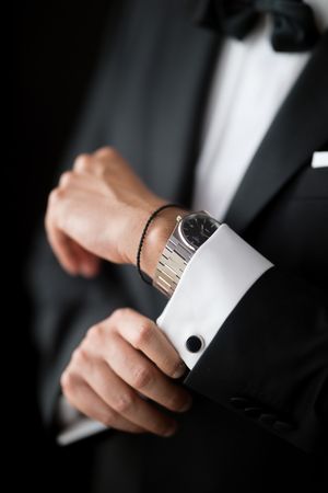 Close-up of hands adjusting silver cufflink and watch while wearing black formal tuxedo suit.