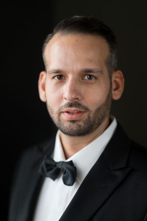 Professional headshot portrait of someone wearing a black tuxedo and bowtie against a dark background.
