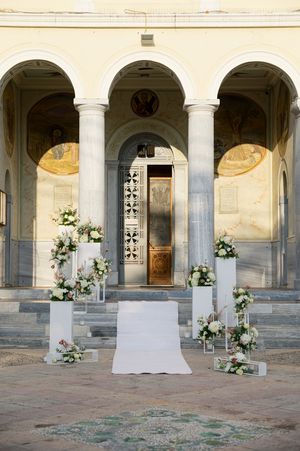 Elegant white floral arrangements decorate a classical arched colonnade with terracotta flooring and faded frescoes.