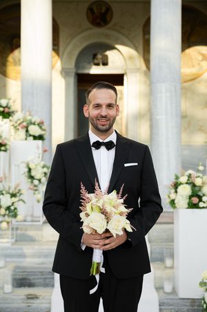 A groomsman in a black tuxedo and bowtie holding a delicate bouquet of white and blush flowers against white columns.