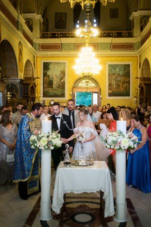 Wedding guests gather around elegant white floral arrangements and decor in an ornate ballroom with crystal chandeliers.