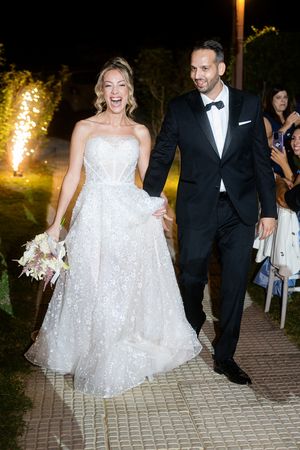 A bride in a sparkling white gown and groom in classic tuxedo walk together joyfully at an evening wedding celebration.