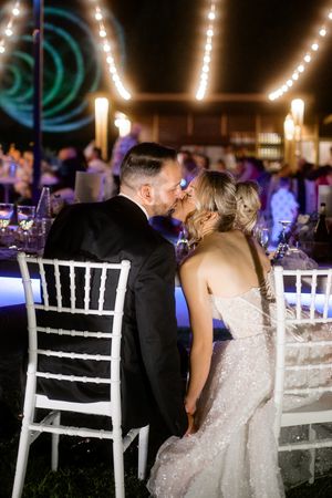A romantic wedding reception moment with twinkling lights and blue uplighting as couple shares an intimate moment at white chairs.