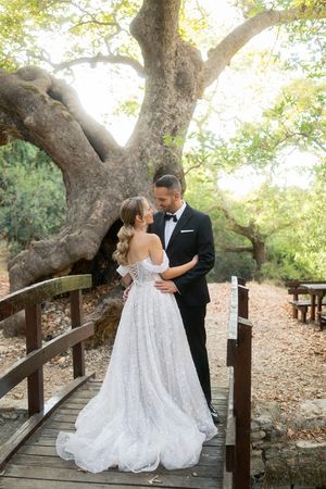 Romantic wedding couple embraces on rustic wooden bridge under majestic oak trees in natural outdoor setting.
