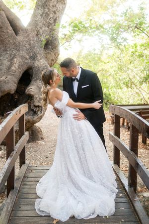 A couple in wedding attire shares romantic moments on a wooden bridge surrounded by large trees.