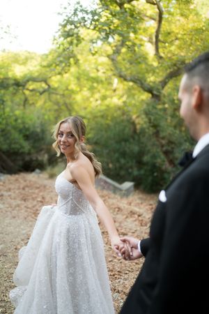A bride in a sparkly white gown dances with her groom on a wooded path during their wedding celebration.