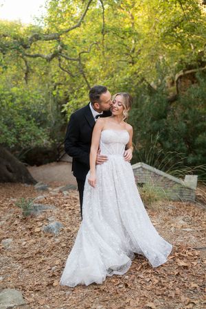 A couple in wedding attire shares romantic moments in a wooded outdoor setting with fallen autumn leaves.