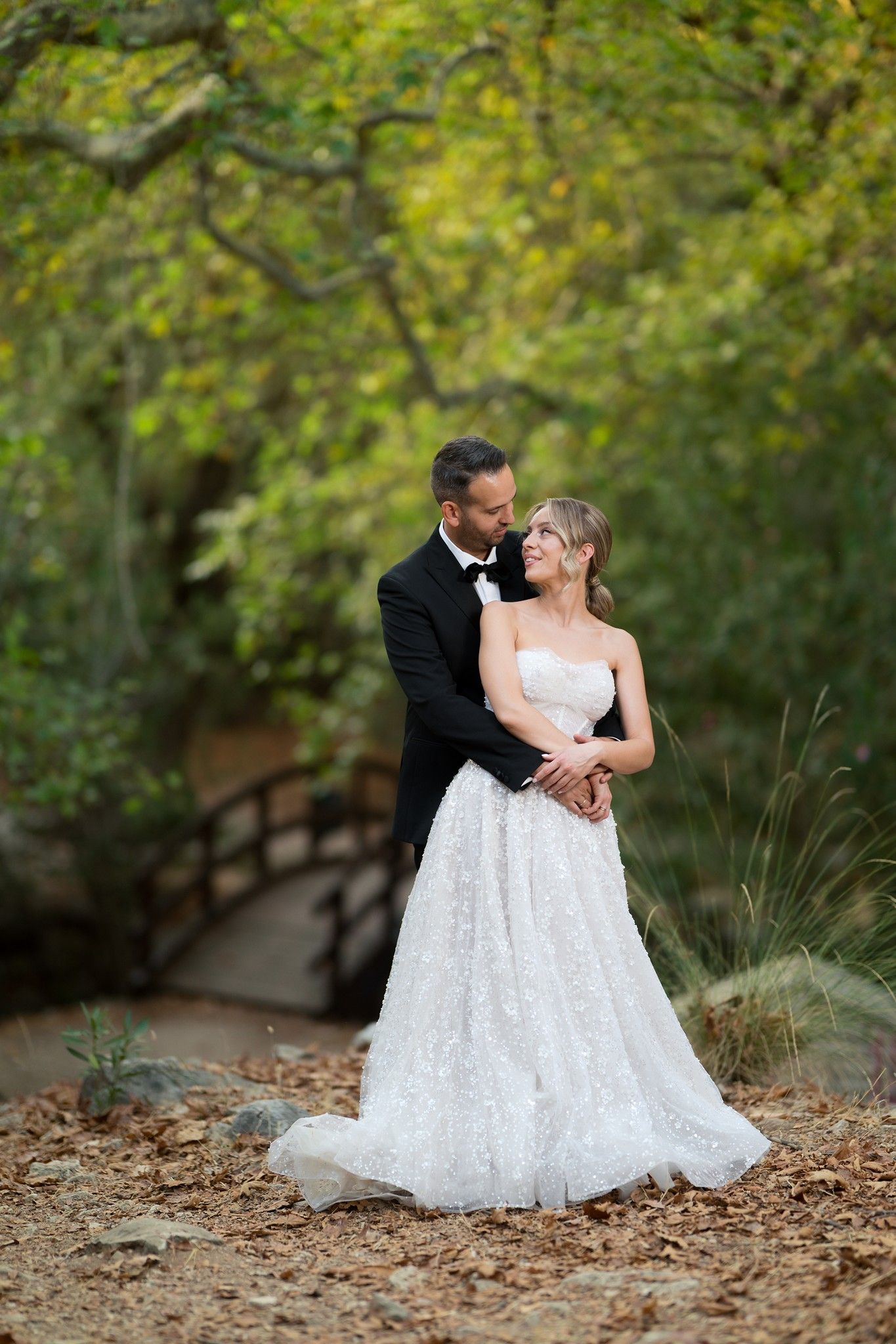 A romantic wedding couple embraces on a woodland path with autumn foliage and a bridge in the background.