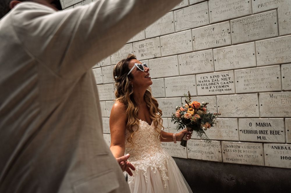 A bride in a white lace wedding dress holds a bouquet while walking along a white brick wall in natural sunlight.