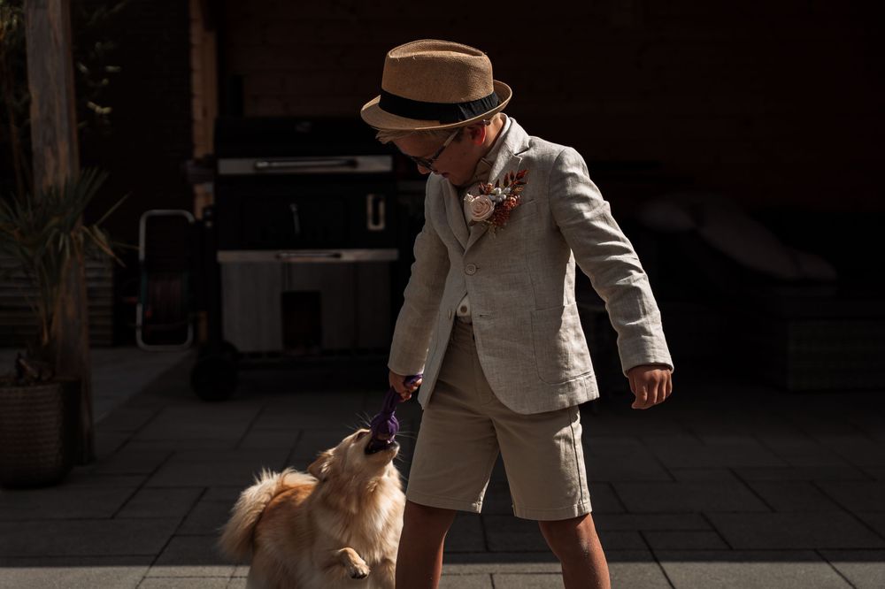 A person in a beige suit and fedora walks with a small fluffy dog on a sunny day.