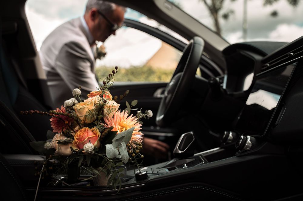 A floral bouquet with orange and peach blooms sits on a black leather car seat while someone enters the vehicle.