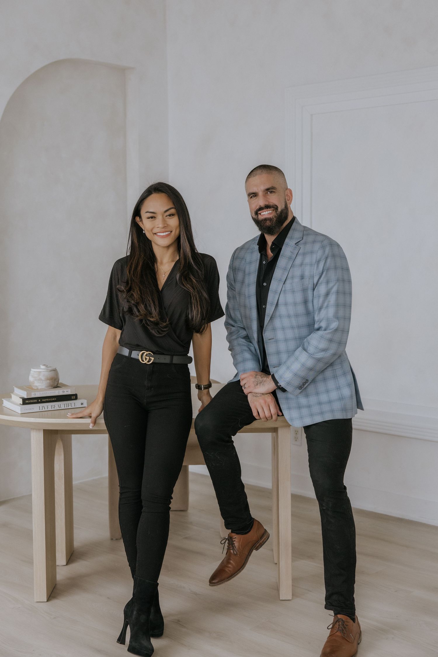 Professional couple poses together in a minimalist studio setting with white walls and wooden furniture.