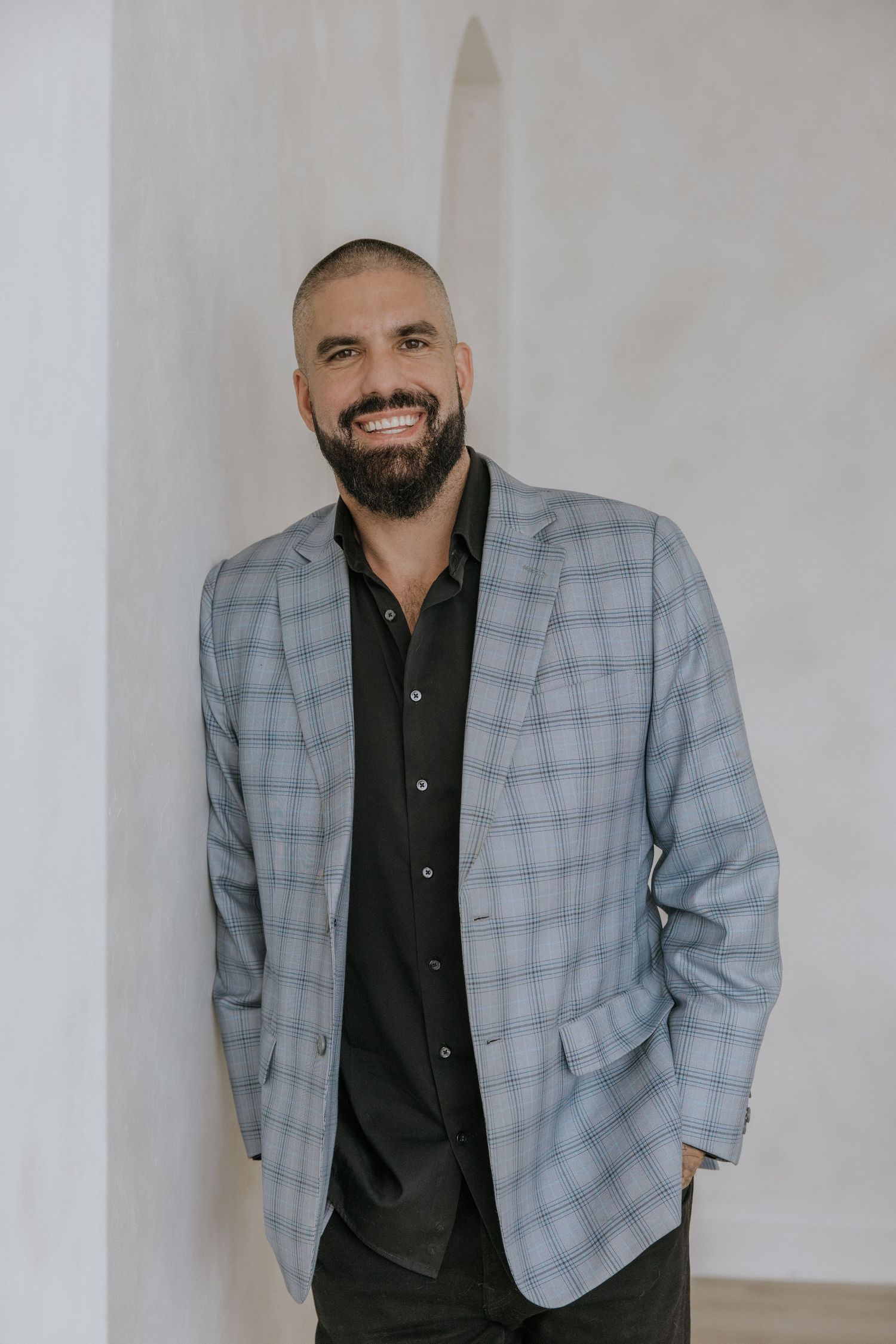 Professional studio portrait showing someone wearing a gray plaid blazer with a black button-up shirt against a white wall.