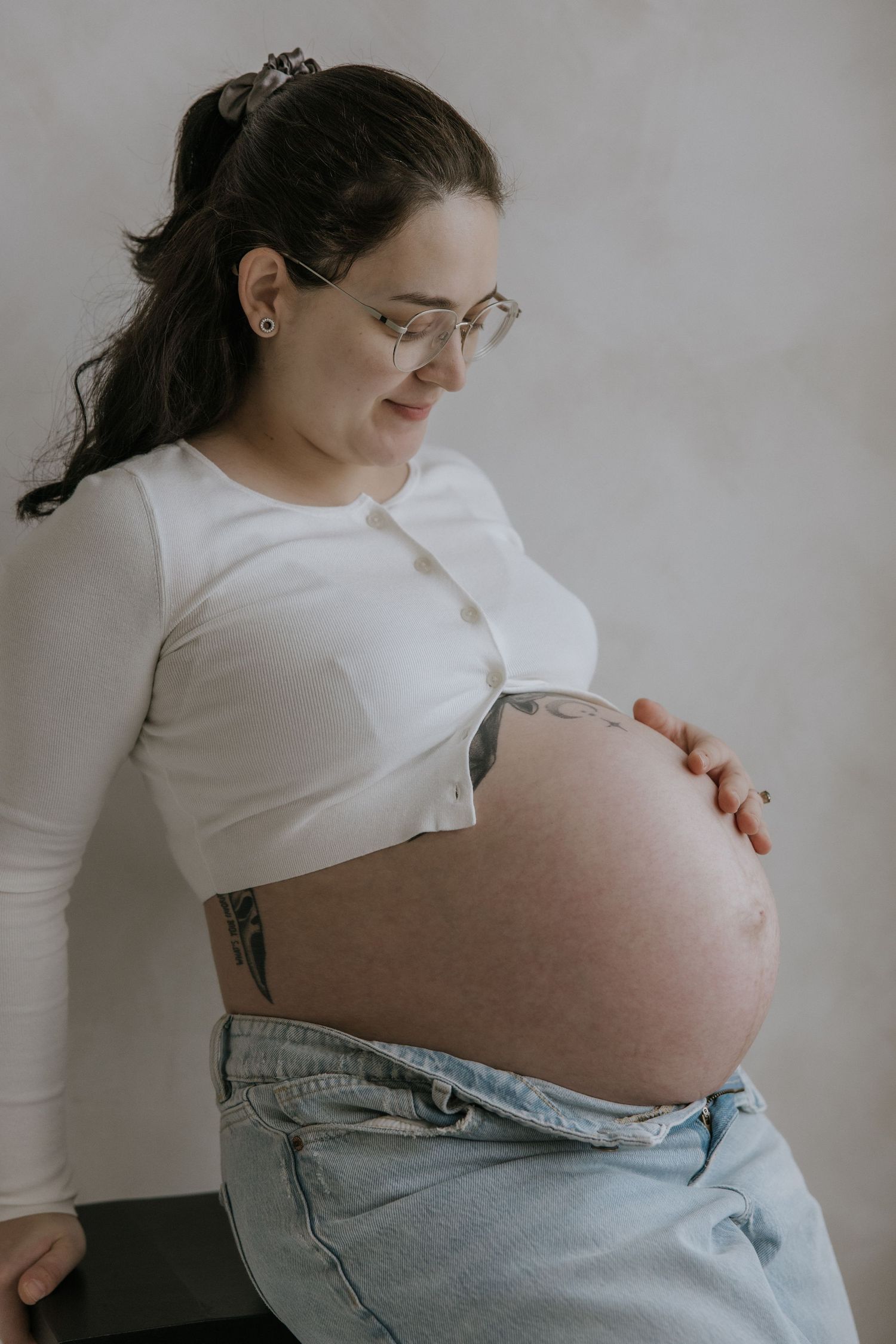 A series of maternity photos showing a pregnant person in a white shirt and jeans touching their belly against a light wall.