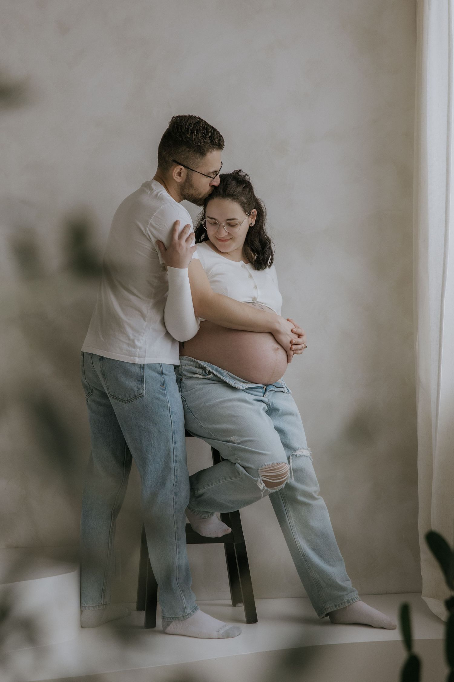 A tender maternity photo session showing a couple in casual attire sharing an intimate moment on a wooden chair.