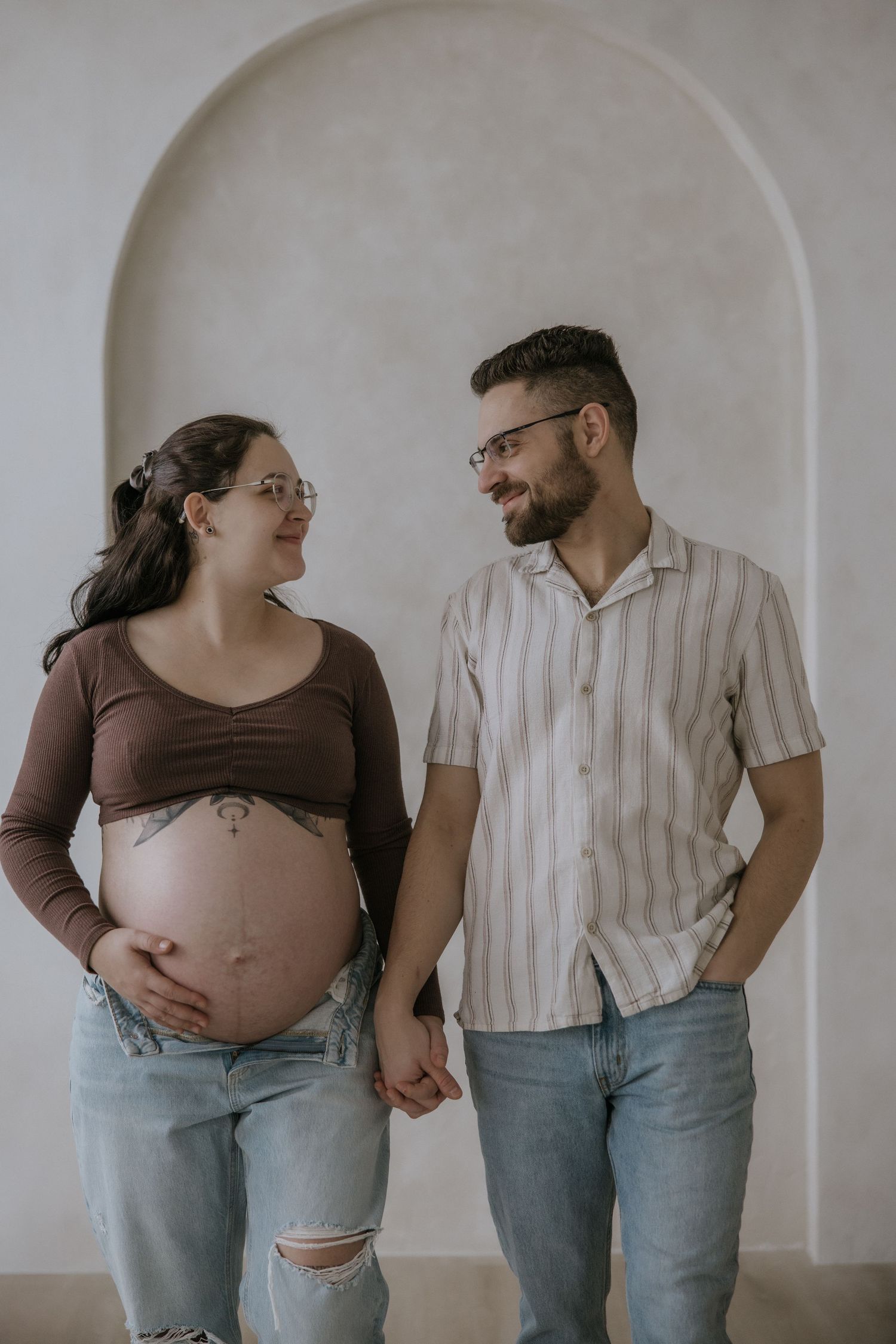 A couple in casual attire share intimate moments while documenting a pregnancy journey against a white wall.