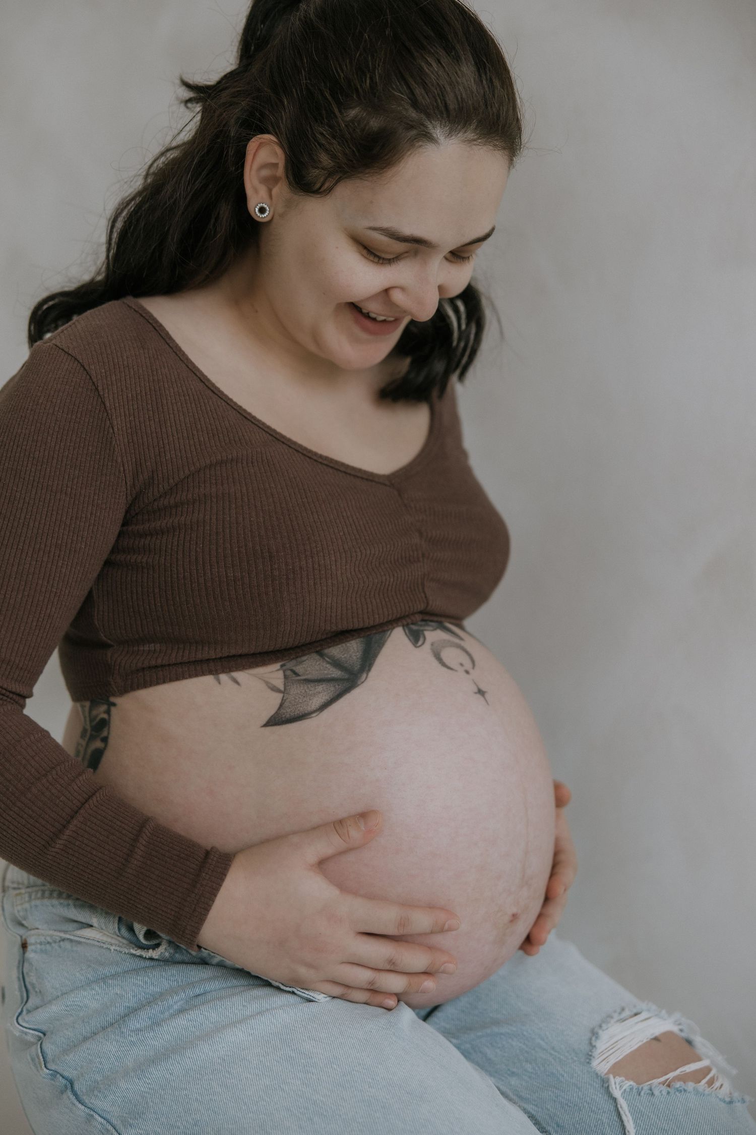 A pregnant person wearing a brown sweater and jeans lovingly cradles their belly while smiling in a series of photos.