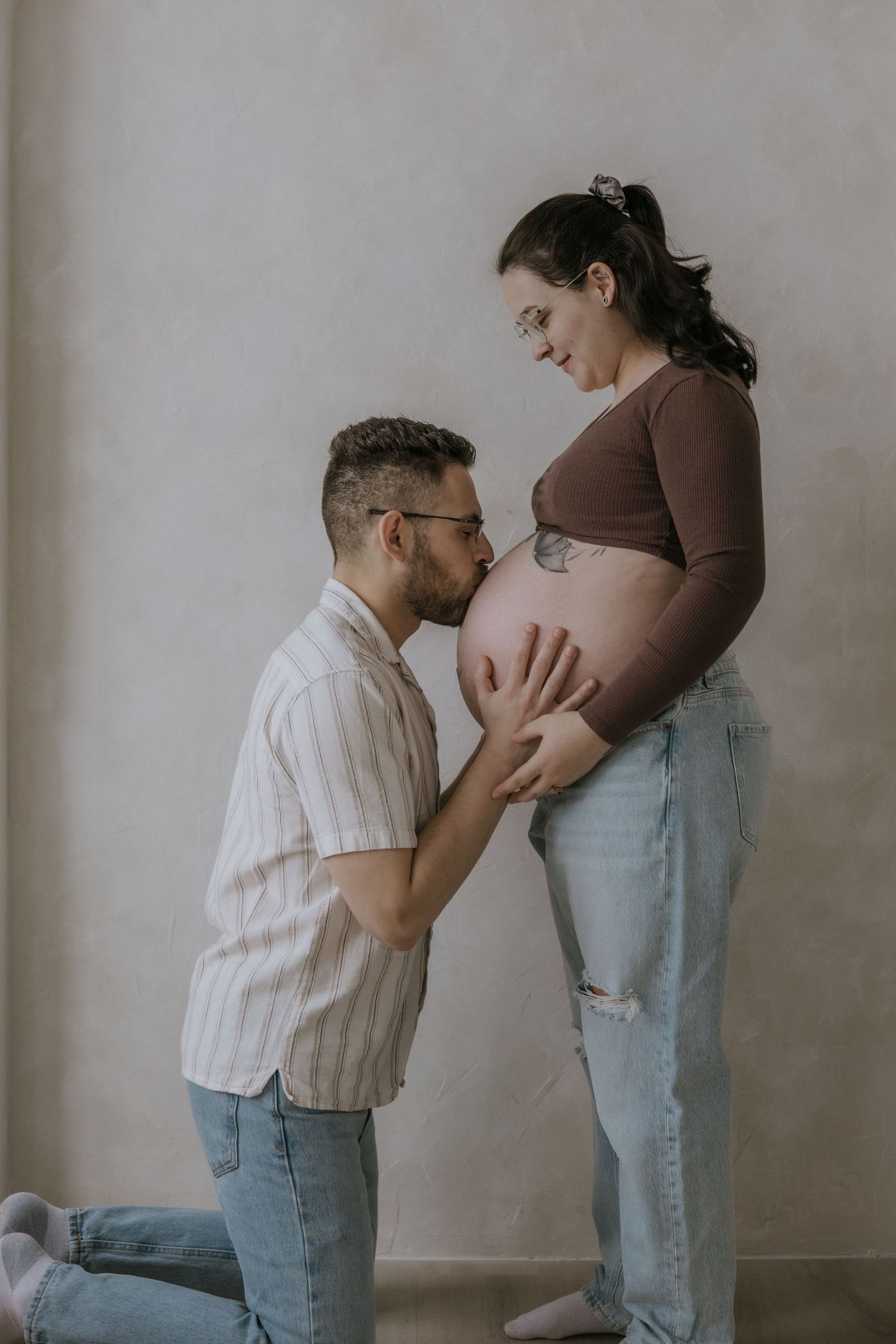 Sequence of intimate maternity photos showing couple embracing with pregnant belly in casual attire against neutral wall.