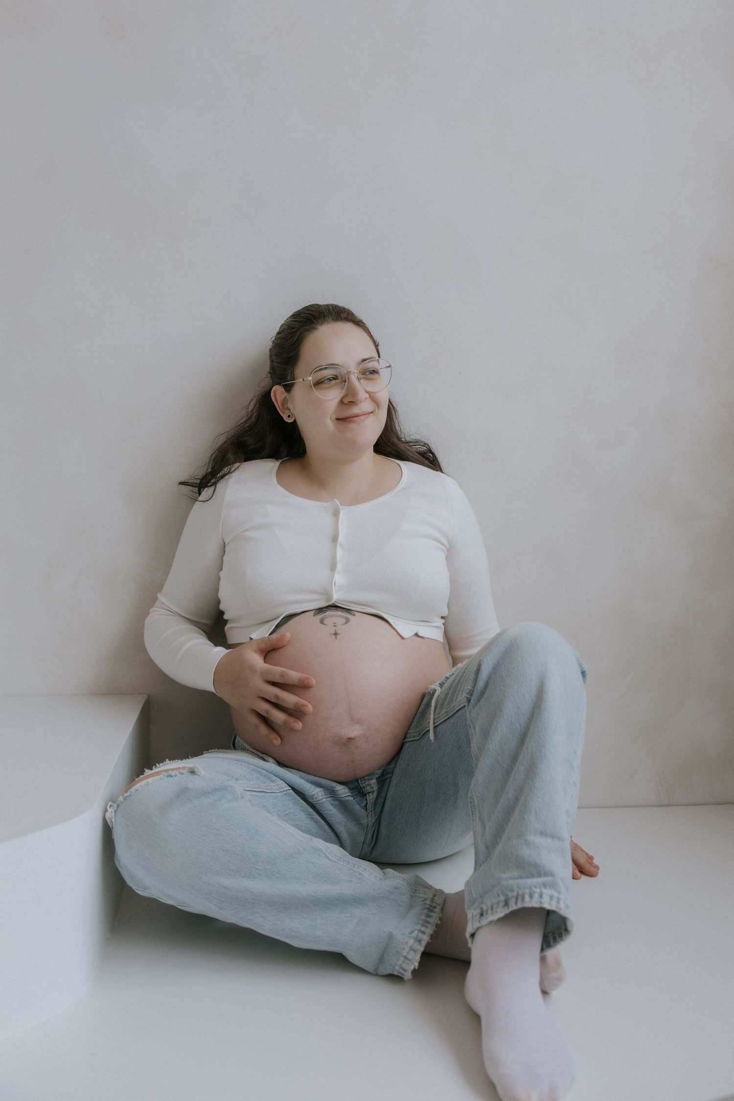 Expectant mother in white top and jeans sits comfortably against light wall while cradling her pregnant belly.