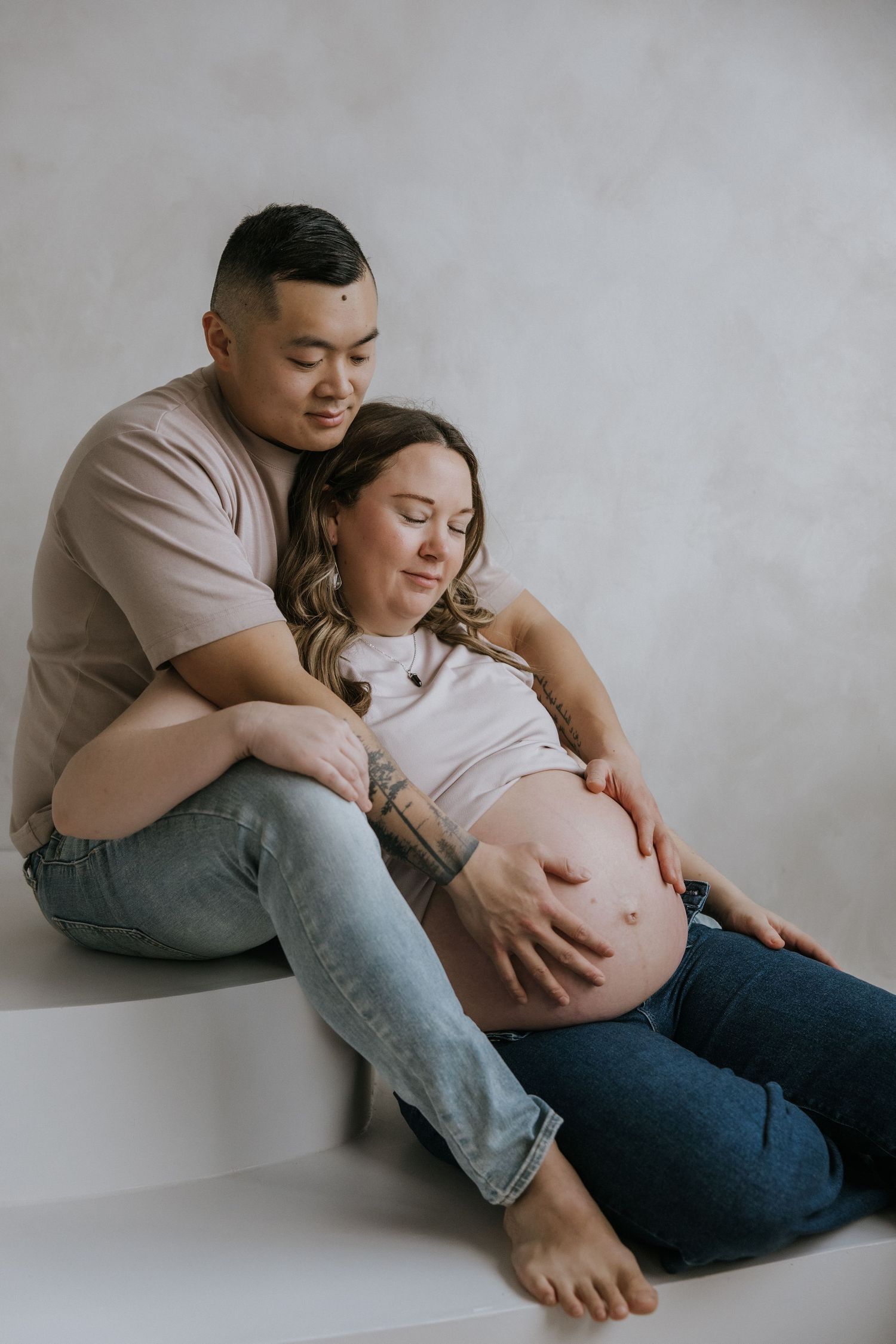 Intimate maternity photo series showing couple in casual clothing against neutral background during pregnancy photoshoot.
