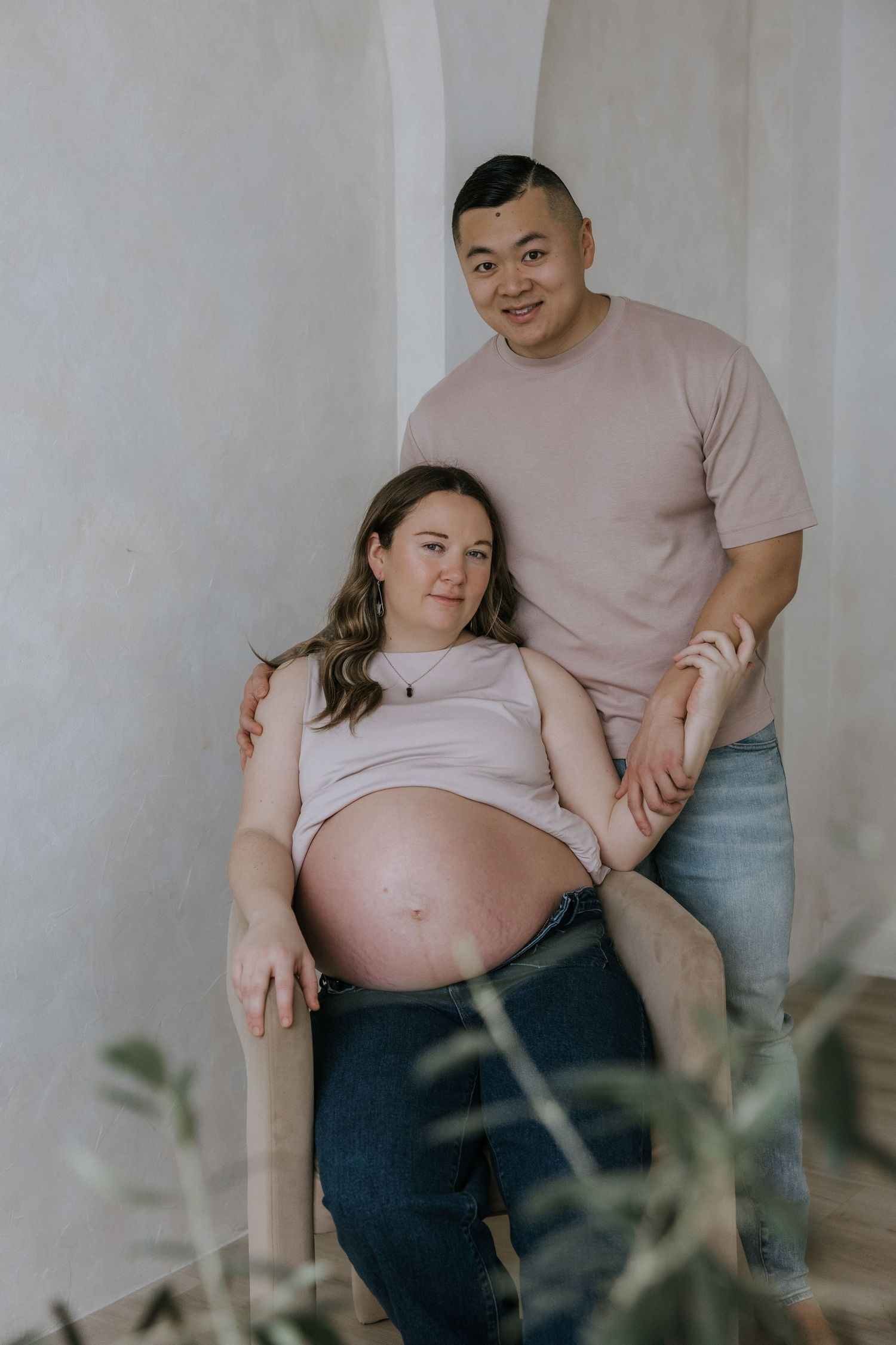 A maternity photo series showing a pregnant woman in light clothing sitting with a partner against a white wall.