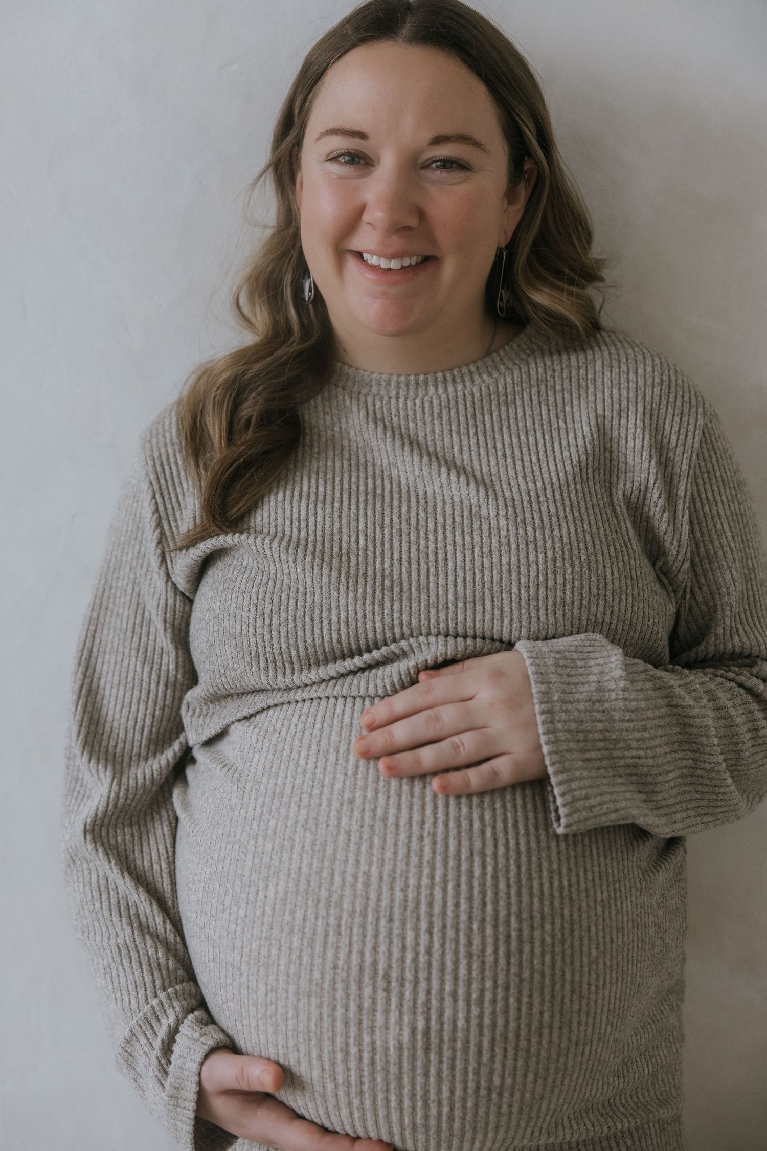 Pregnant woman in gray sweater shows progression of belly touches and smiles against neutral wall background.